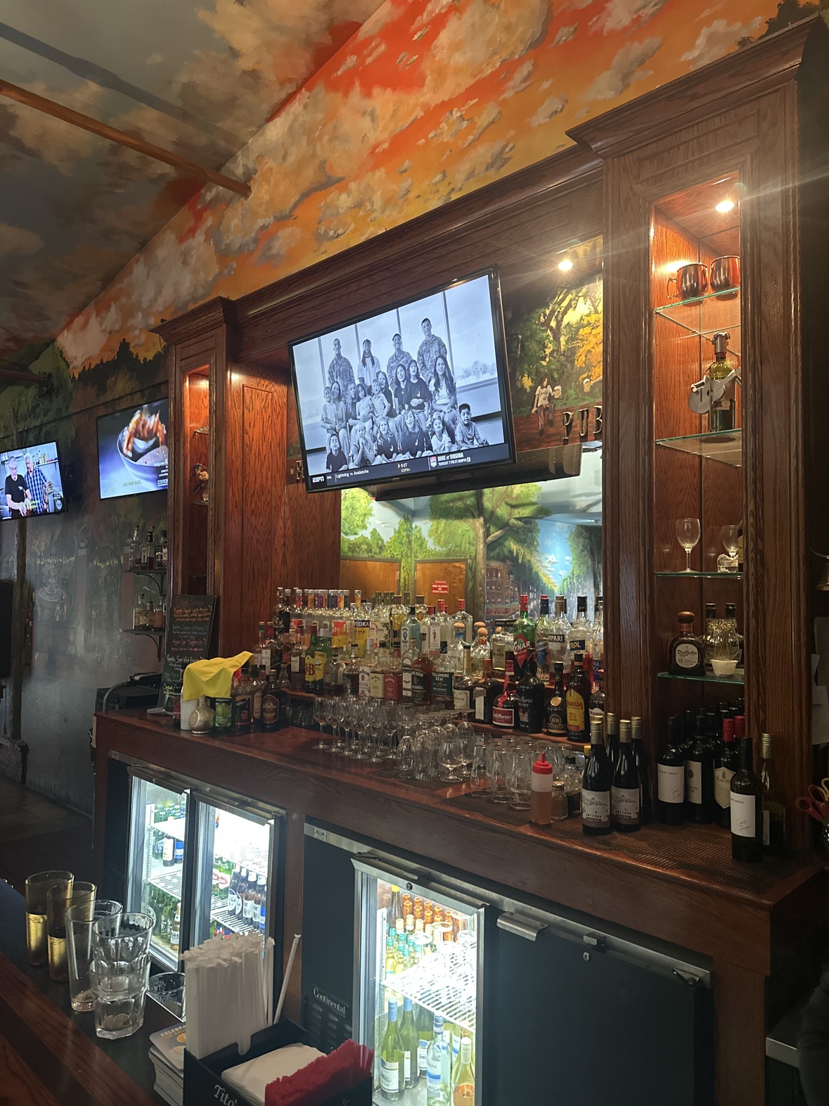 The bar at Ferraro's — a full bourbon shelf and rows of bottles under the painted sunset ceiling, with a mural behind the mirror